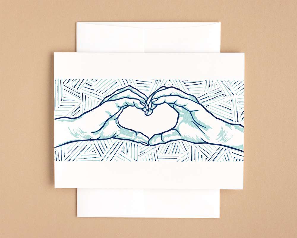 A horizontal white card depicts a blockprinted pair of hands making a heart shape. The design is done in navy and light blue. The card sits on a white envelope, which lies on a brown backdrop.